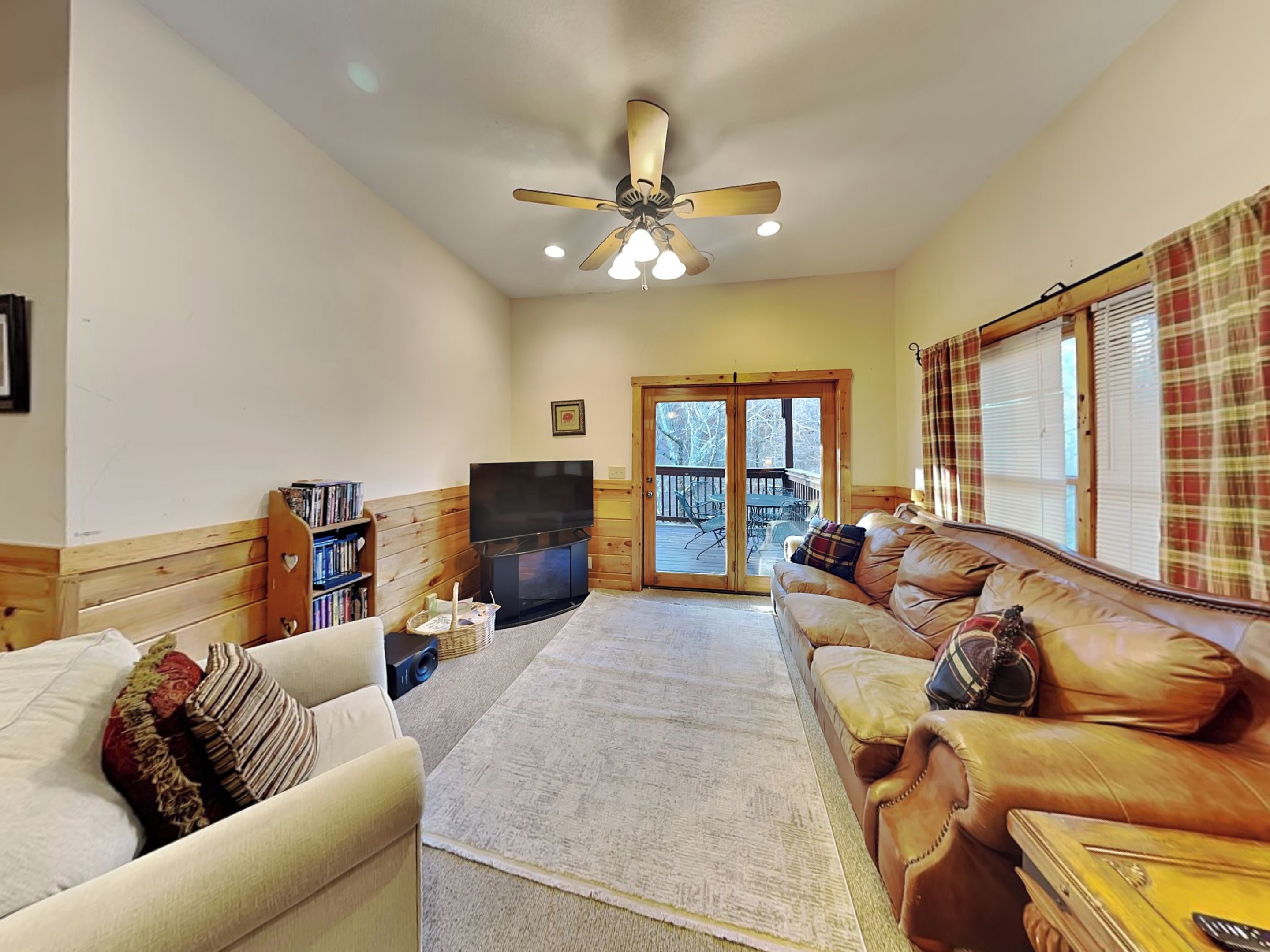 A cozy living room with a sofa, a television, a bookshelf, and a large window with blinds.