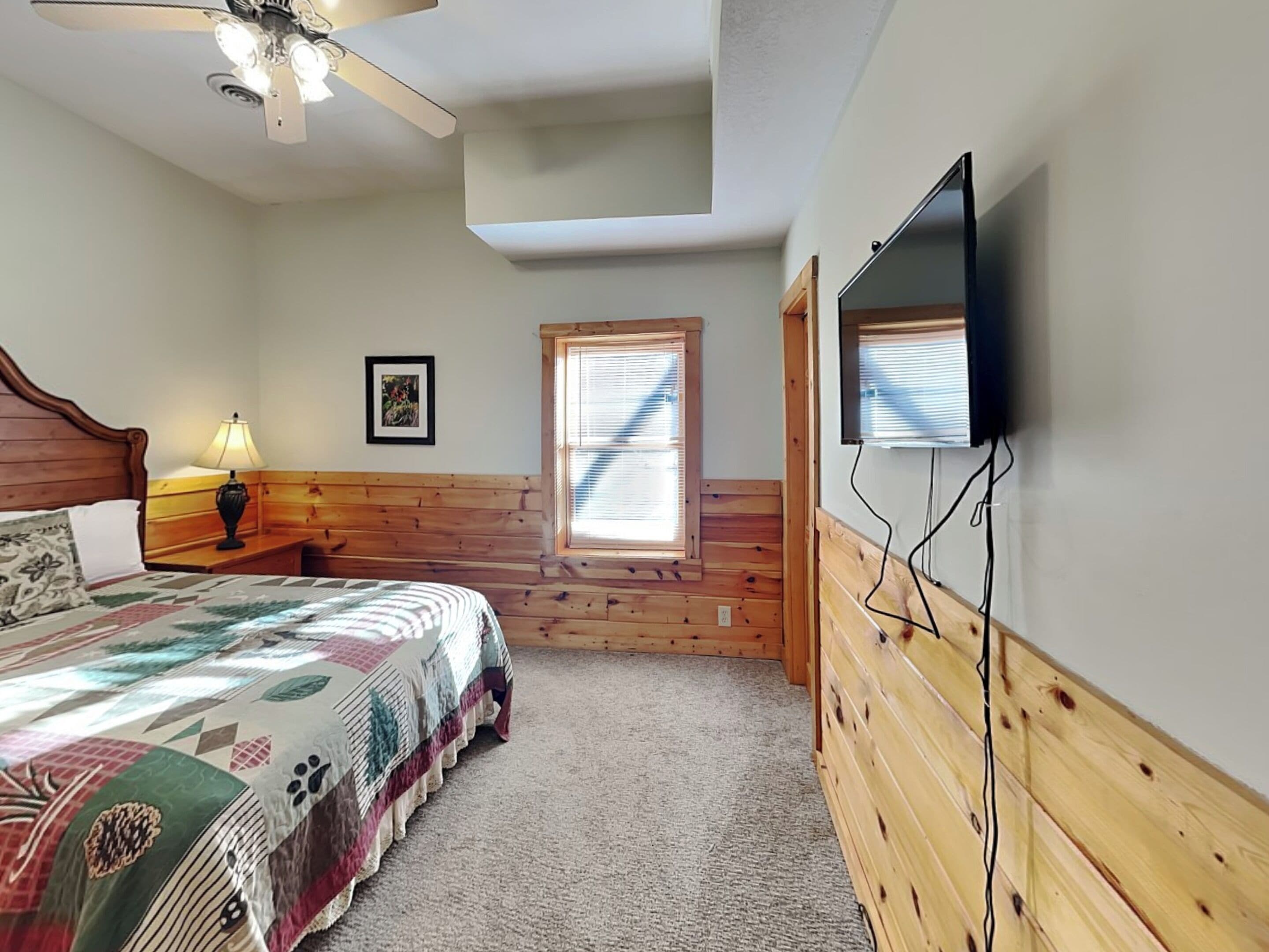A bedroom with a bed, a ceiling fan, a TV mounted on the wall, a window with blinds, and a wooden headboard.
