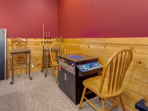 A room with wooden panel walls, a classic arcade machine, and several wooden chairs.