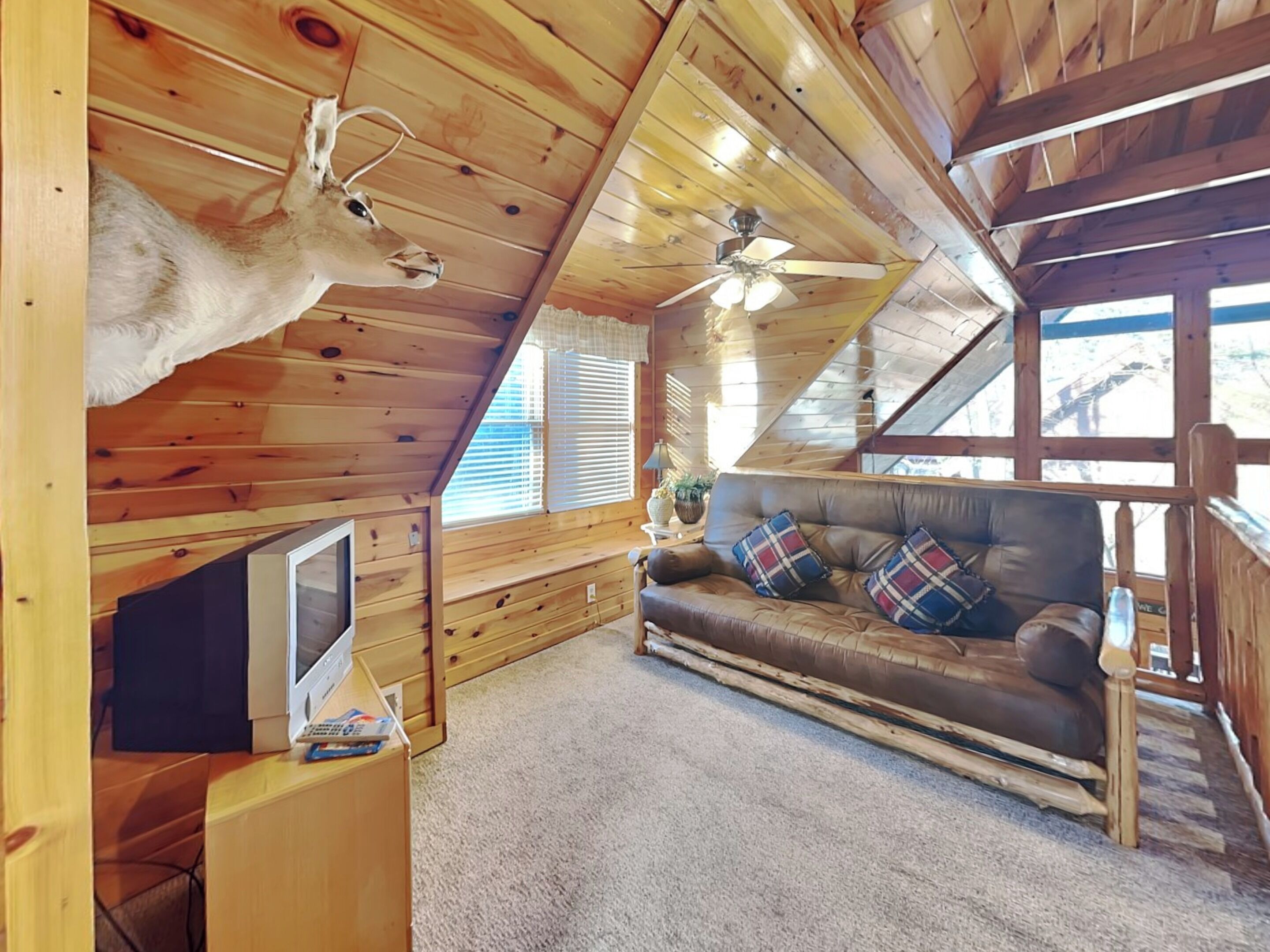 Interior acogedor de una cabaña con techo de madera, un sofá de cuero con cojines escoceses, una cabeza de venado montada y un ventilador de techo.