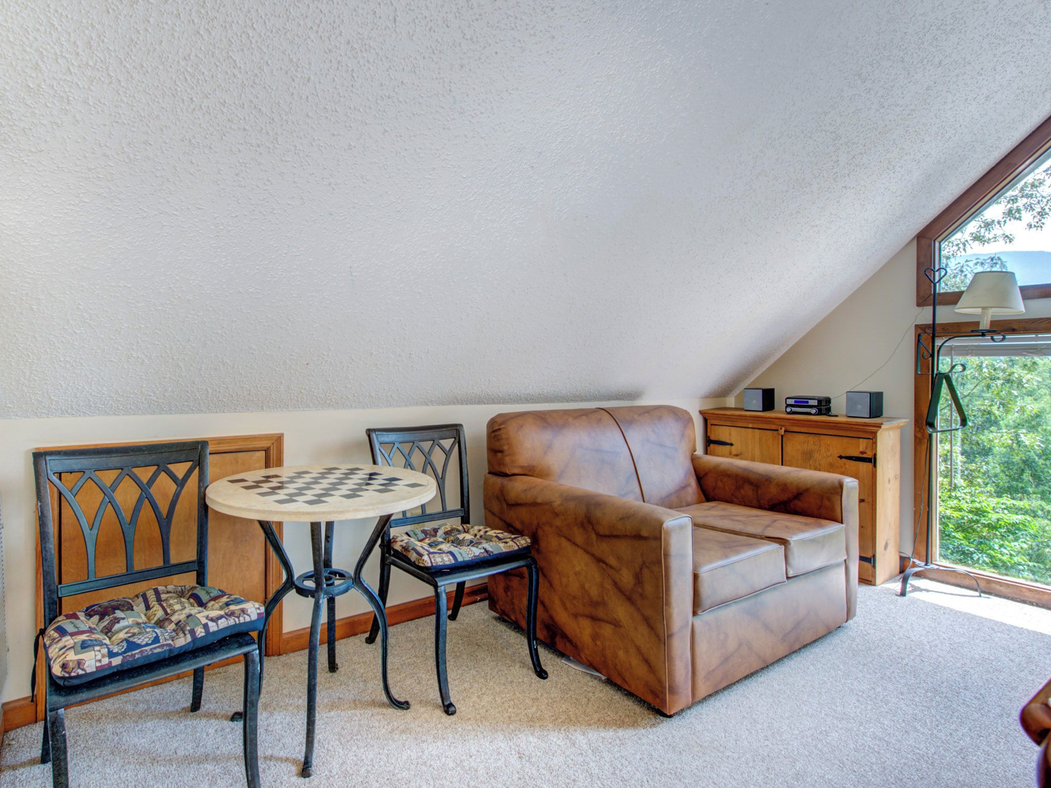 A cozy attic room with a brown leather sofa, a wooden cabinet, and a small round table with a chessboard.
