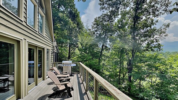 A wooden deck with outdoor furniture, a grill, and a view of a forested area. - Mountain Charm by Jackson Mountain Rentals (Gatlinburg)