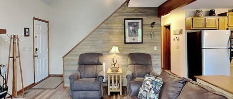 A living room with two armchairs, a sofa, a wooden floor, a small table with a lamp, and a kitchen area with cabinets and a refrigerator.