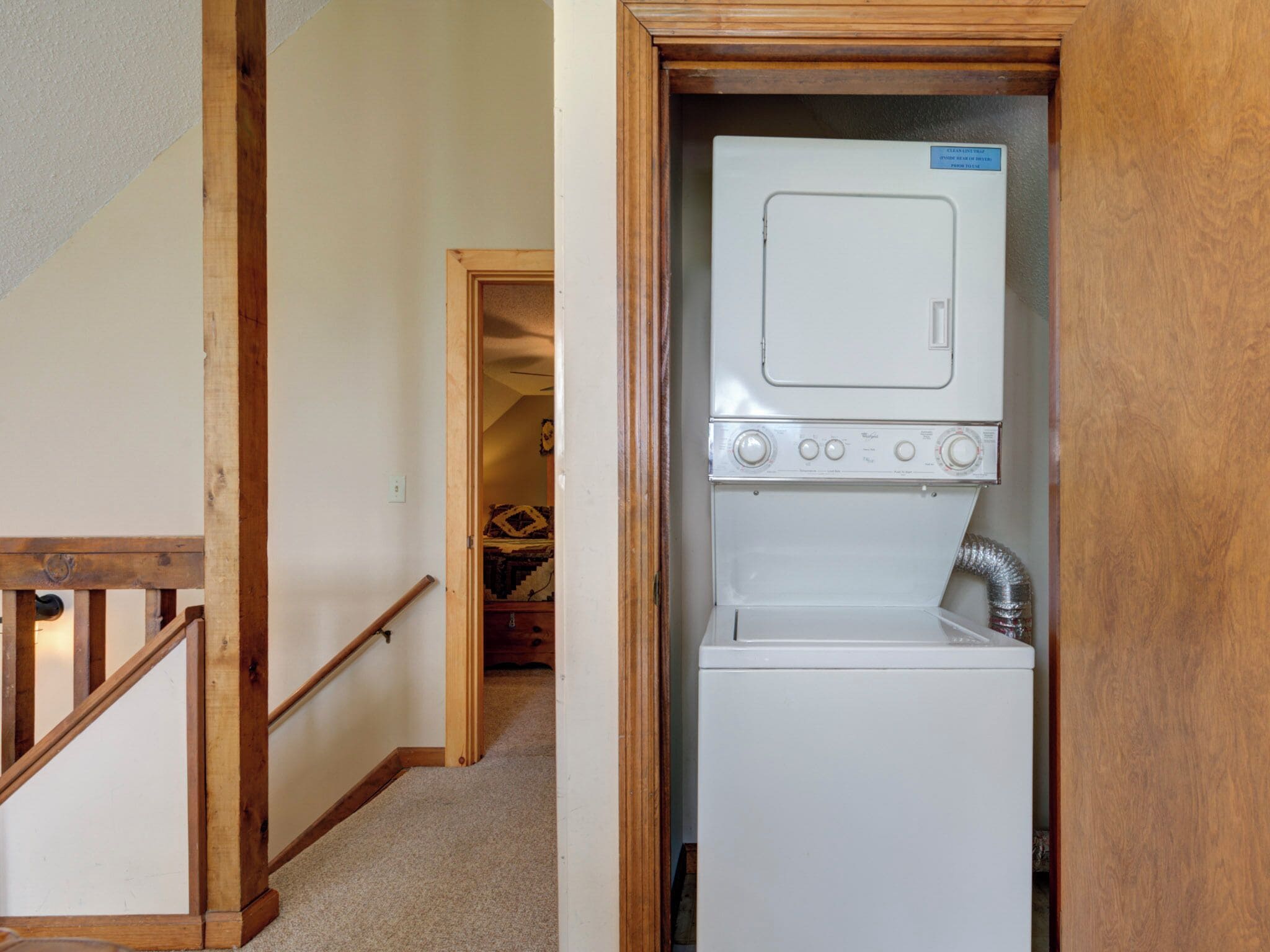 A laundry room with a stacked washer and dryer, wooden staircase, and a carpeted floor.