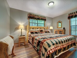 A bedroom with a wooden bed, two bedside lamps, a nightstand, a window with blinds, a mirror, and a fireplace. - Mountain Charm by Jackson Mountain Rentals (Gatlinburg)