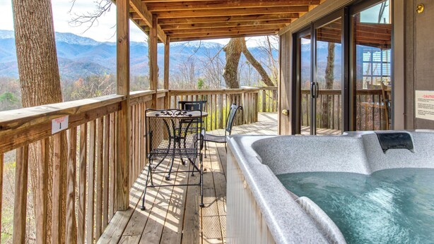 A wooden deck with a hot tub, a table, and chairs, overlooking a mountainous landscape. - Mountain Treasure by Jackson Mountain Rentals (Gatlinburg)