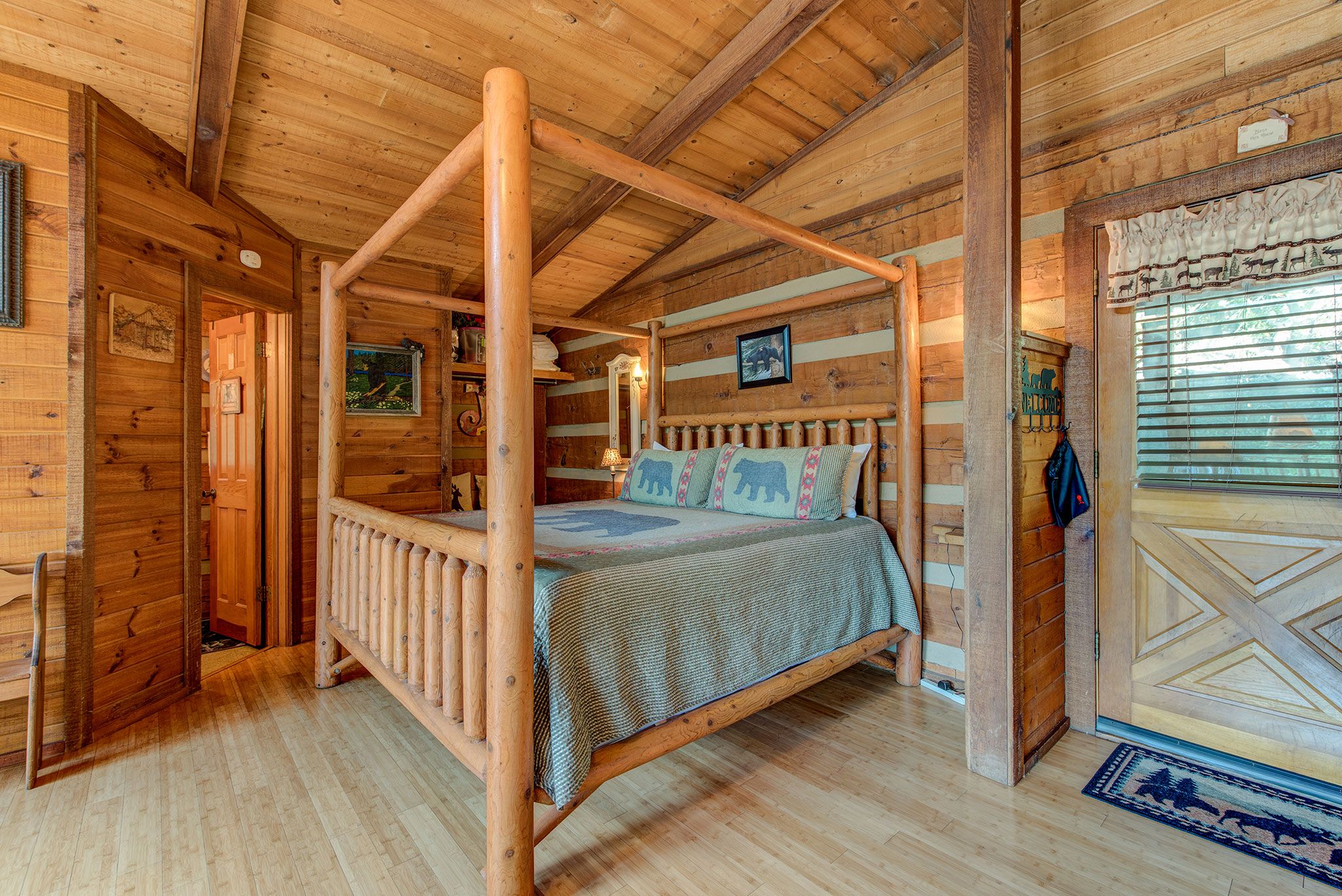 A wooden four-poster bed in a log cabin room with a wooden floor and a door with a geometric pattern.