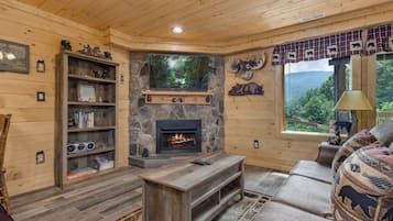 Un interior acogedor de cabaña con chimenea de piedra, estanterías de madera, un sofá y un ventanal con vistas al exterior.