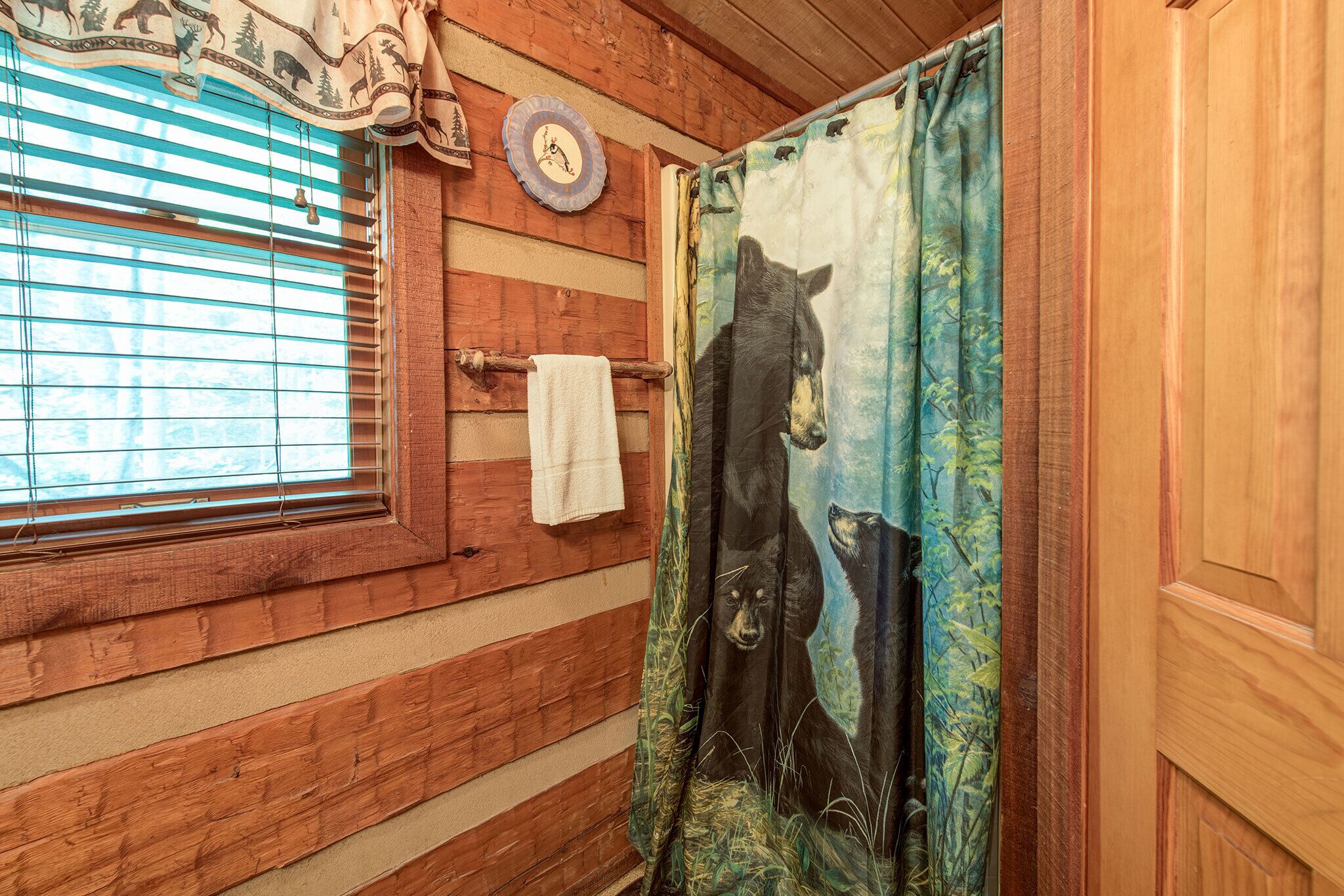 Interior de una cabaña de madera con una ventana, una toalla colgada de la puerta y cortina de ducha con motivos de oso.
