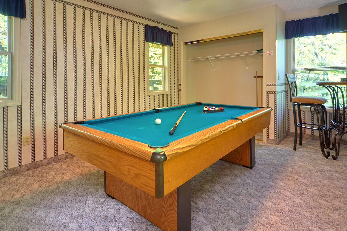 A pool table in a room with striped wallpaper, a window with a view, and a chair.
