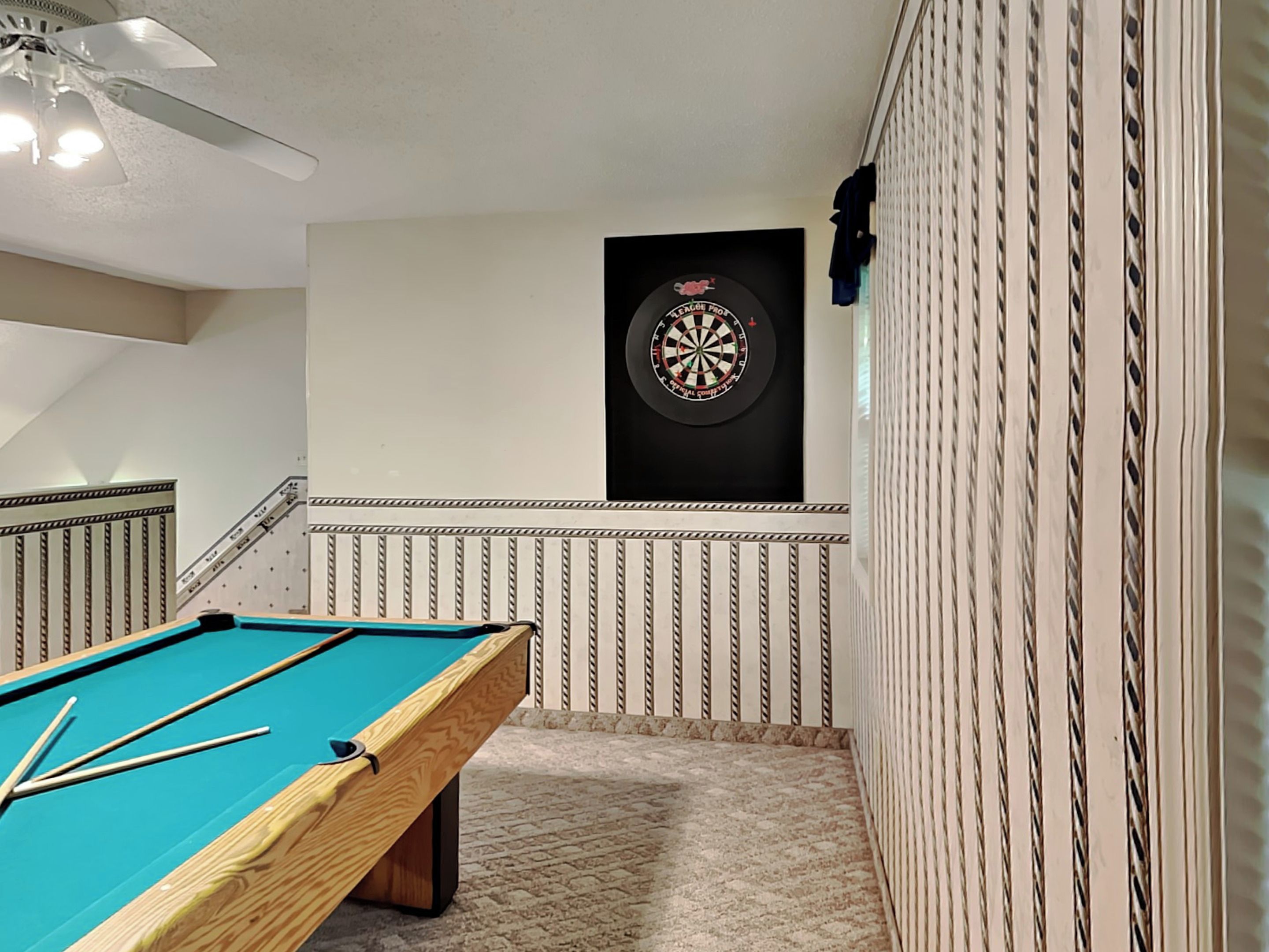 A billiards table with a dartboard in a room with patterned walls and a ceiling fan.