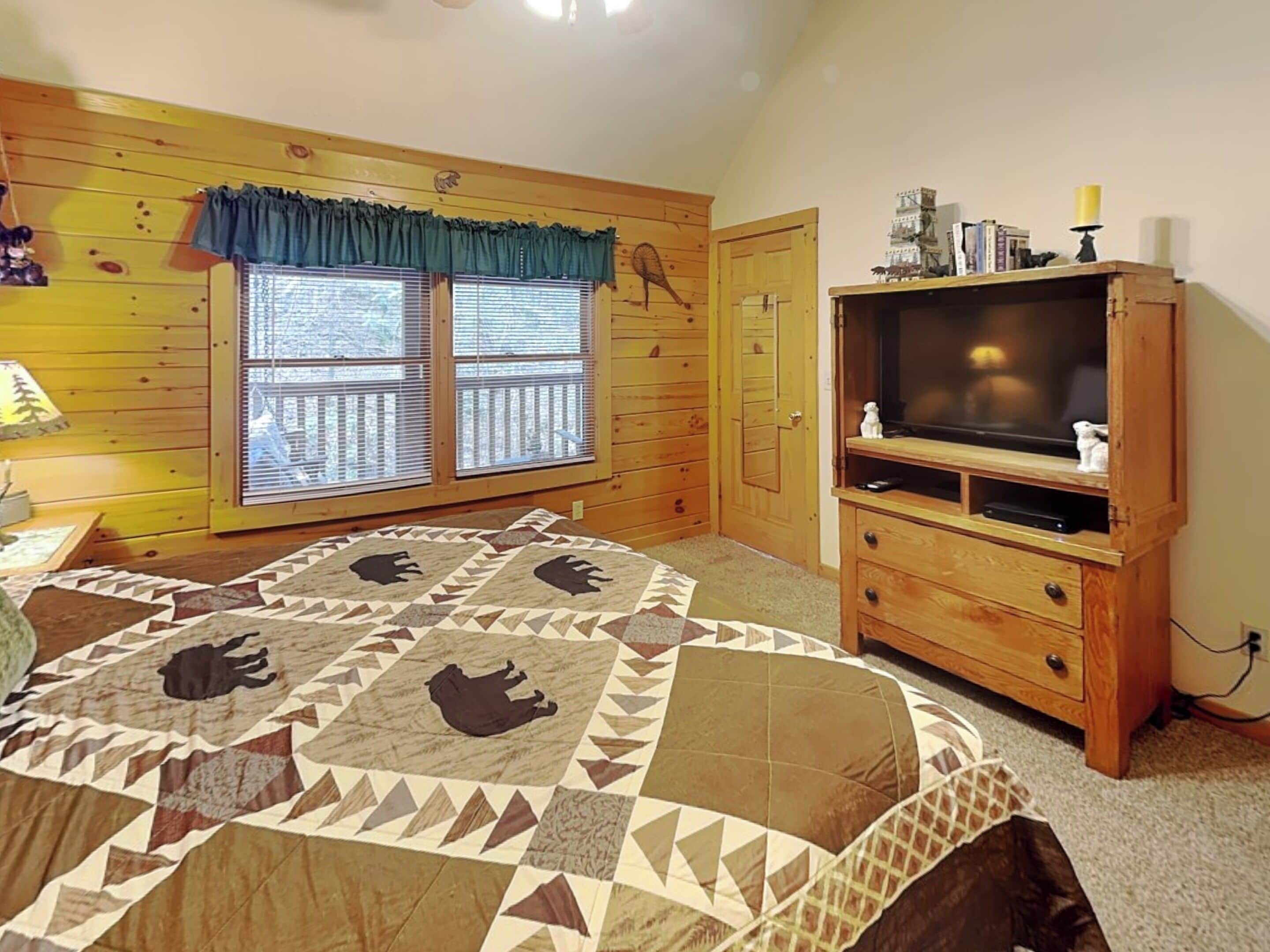 A bedroom with a wooden floor, a bed with a patterned quilt, a television on a wooden stand, and a window with blinds.