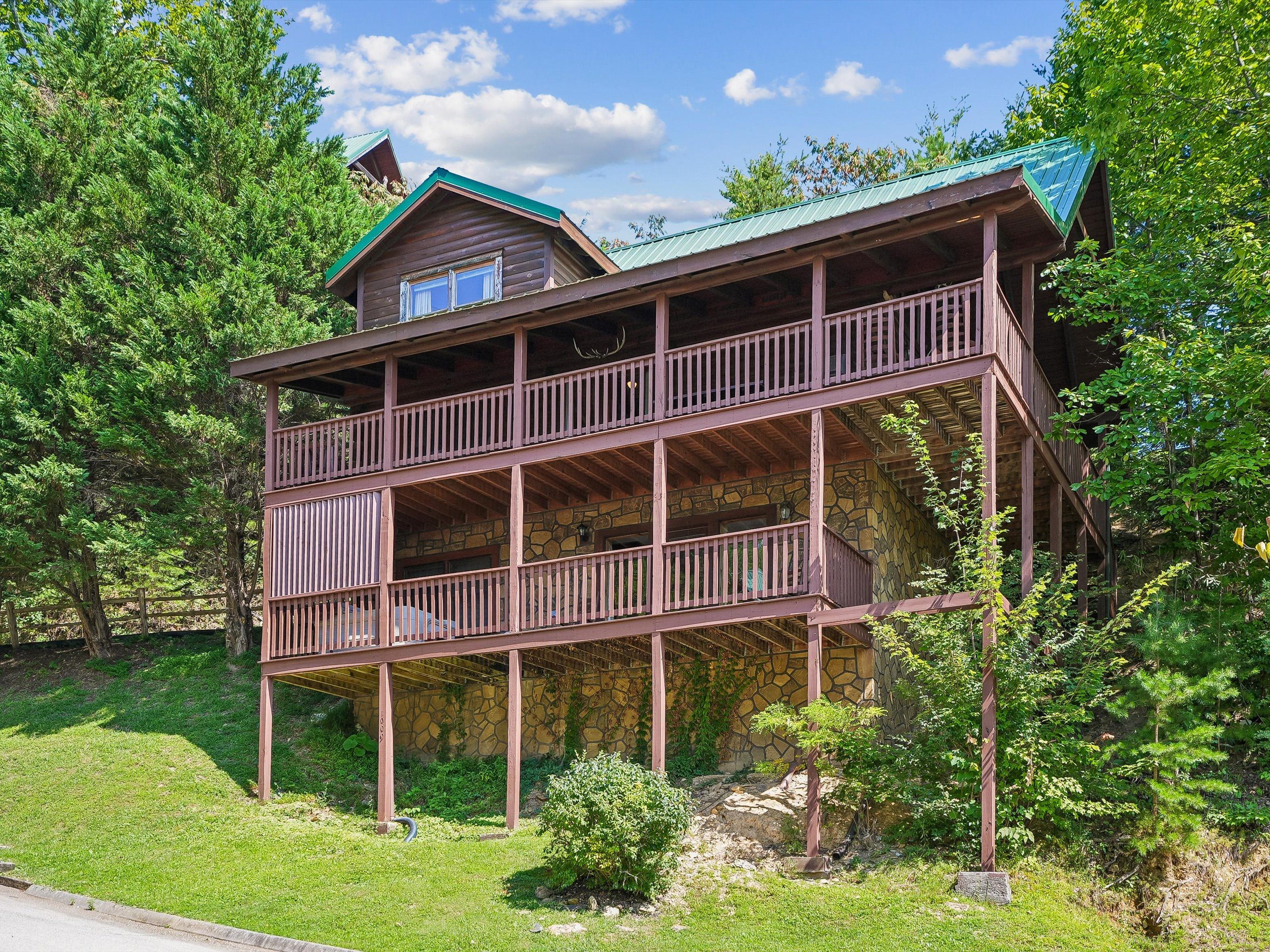 A two-story wooden cabin with a balcony, surrounded by trees and a stone wall.