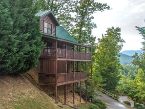 Une cabane dotée d’un balcon à deux étages, entourée d’une forêt dense et offrant une vue sur des collines lointaines.