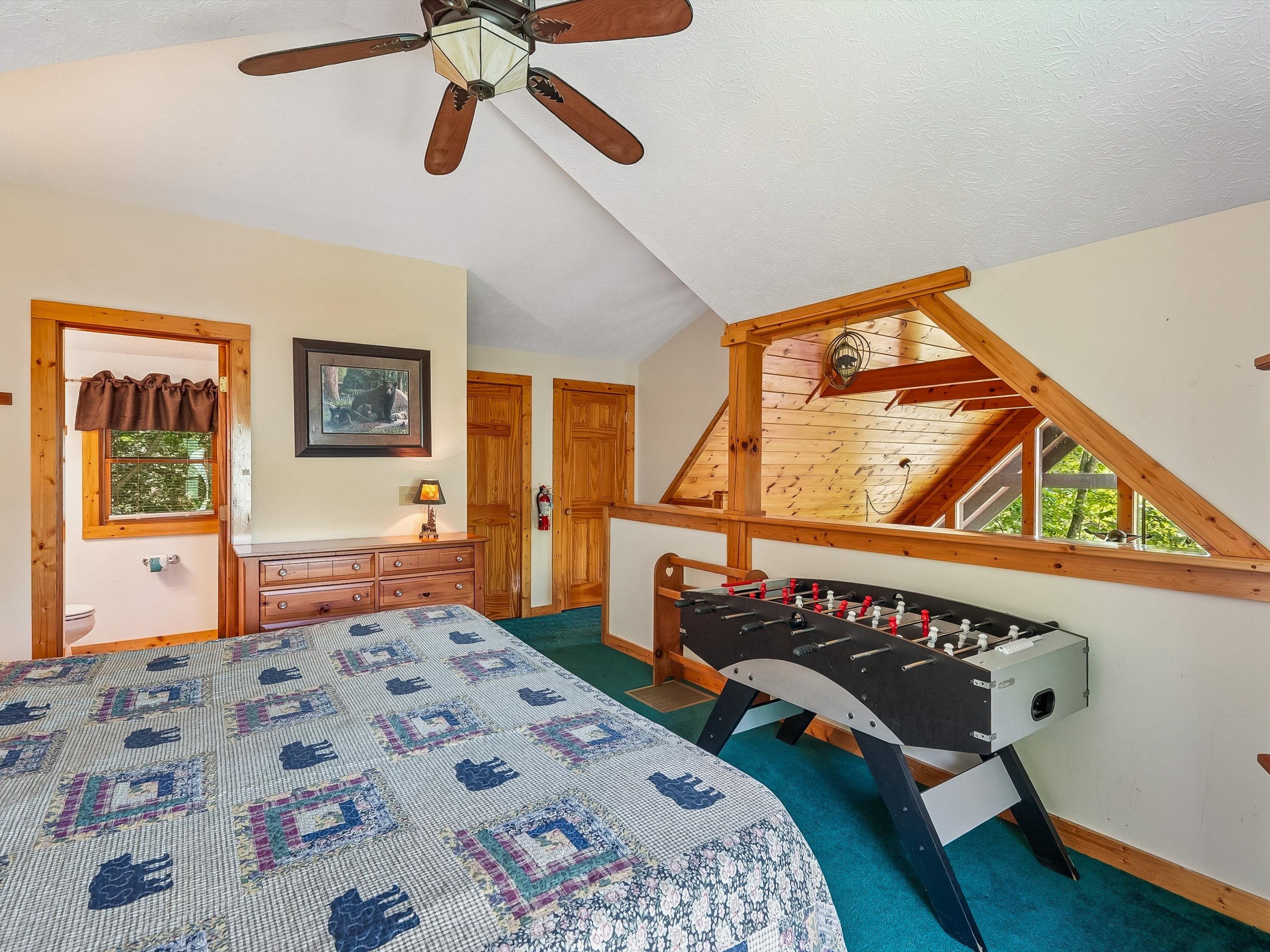A bedroom with a bed, a wooden ceiling fan, a window with a view, and a foosball table.
