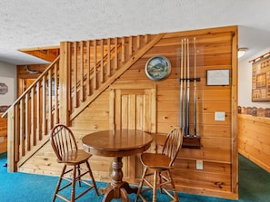 A wooden interior with a round table and two chairs, a clock, and a pool table with cues. - Komfy Kozy by Jackson Mountain Rentals (Gatlinburg)
