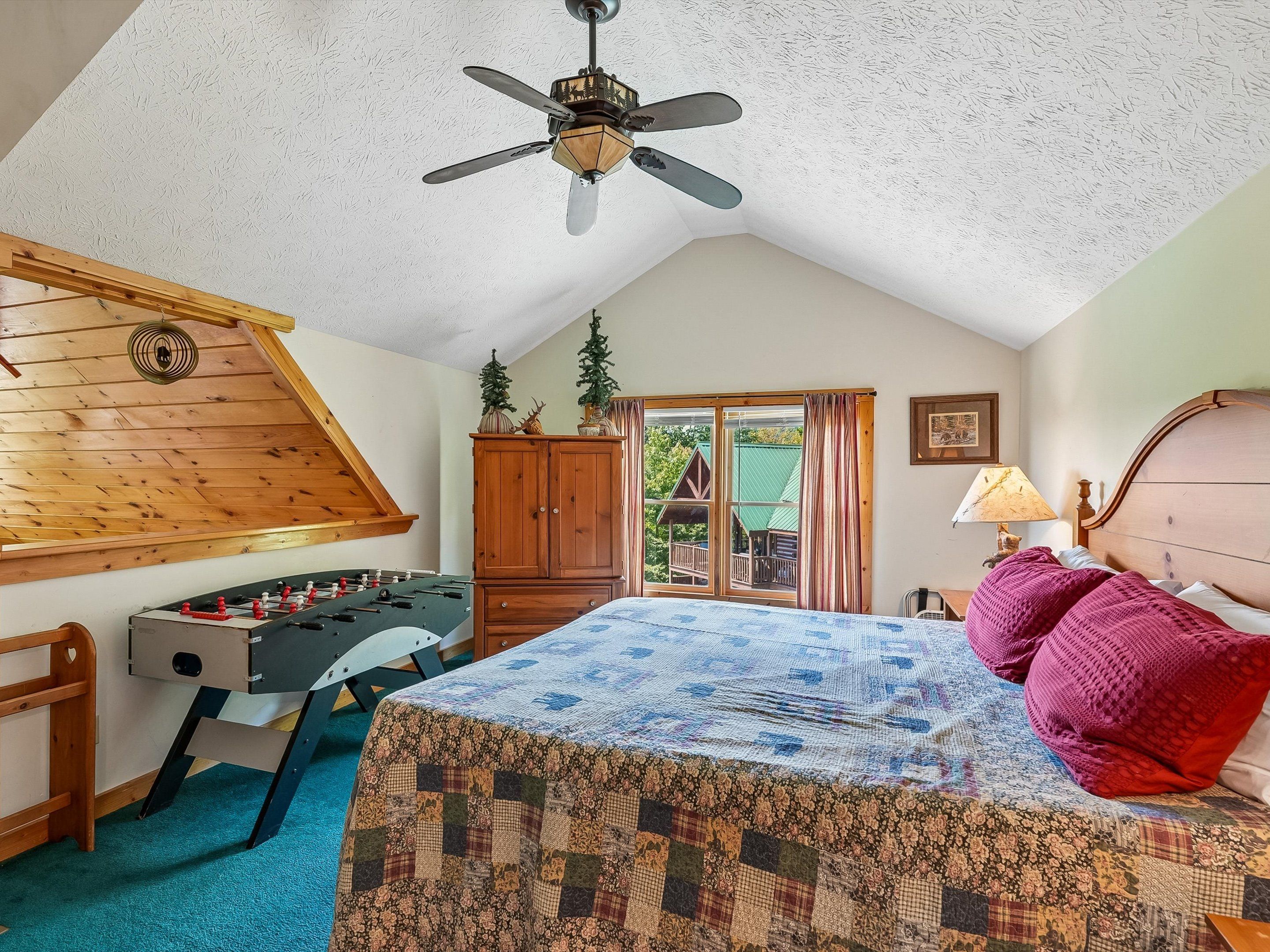 Quarto com cama, ventoinha no teto, guarda-roupa de madeira, janela com cortinas e vista para uma casa lá fora.