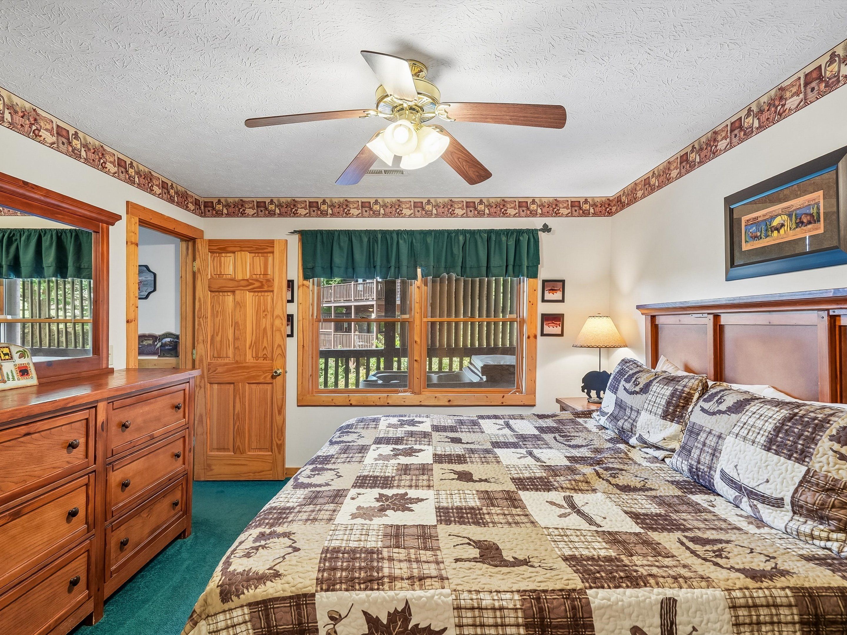 A bedroom with a bed, a ceiling fan, a window with a view, a dresser, and a lamp.