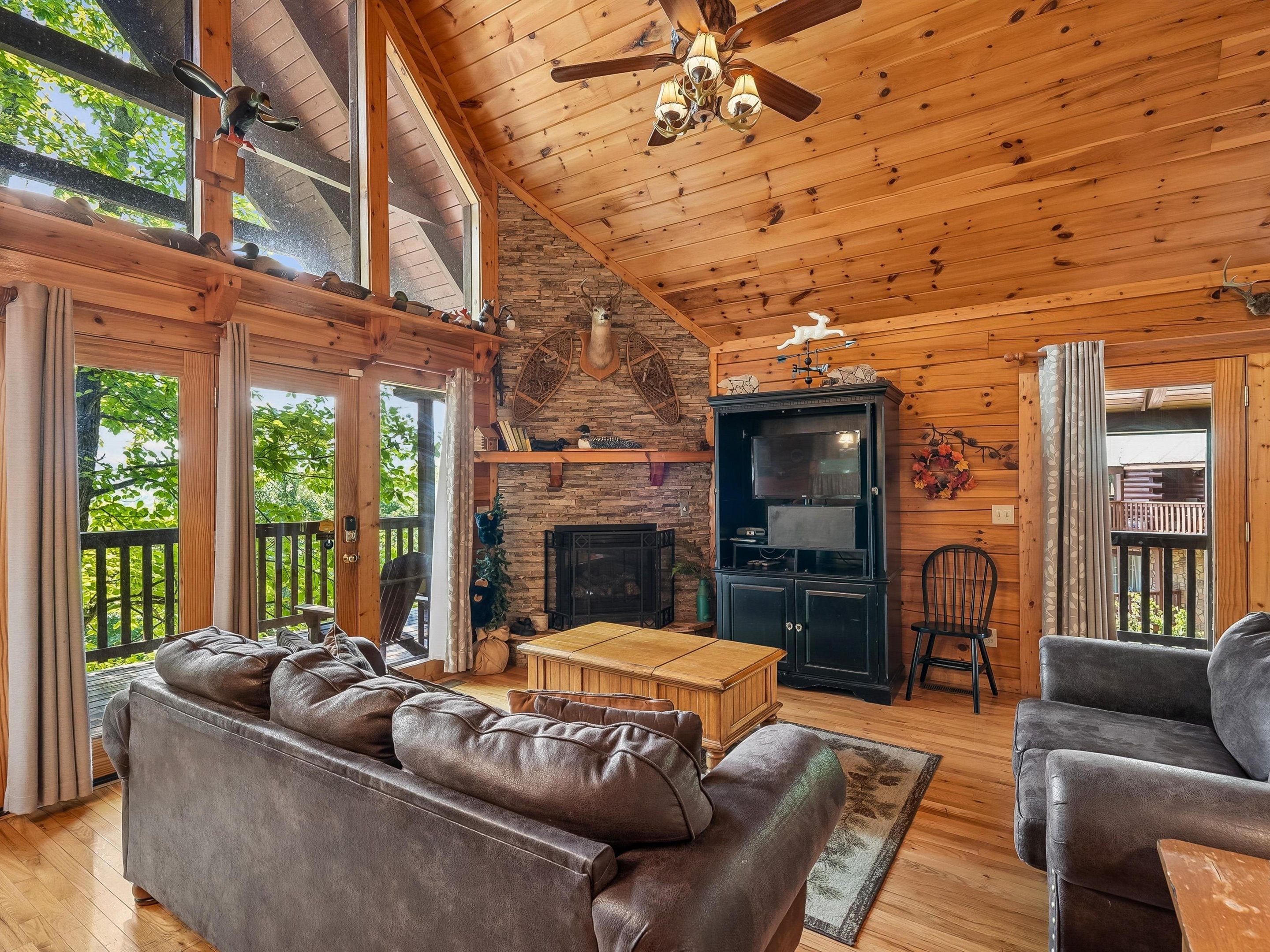 A cozy cabin interior with a fireplace, leather sofa, wooden furniture, and a balcony with a view of trees.