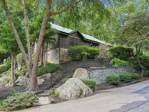 A log cabin with a stone foundation, surrounded by lush greenery and a stone staircase leading to the entrance. - Komfy Kozy by Jackson Mountain Rentals (Gatlinburg)