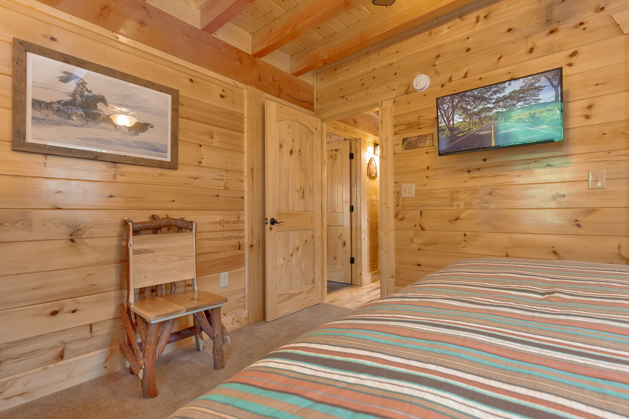Habitación tipo cabaña de madera con una cama, una silla, un televisor y una puerta que da a otra habitación.
