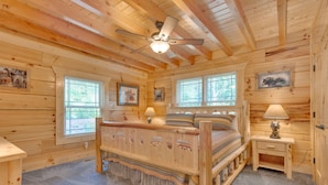 Une chambre dans une cabane en bois, avec un lit, deux fenêtres, un ventilateur de plafond et une table de chevet avec une lampe.