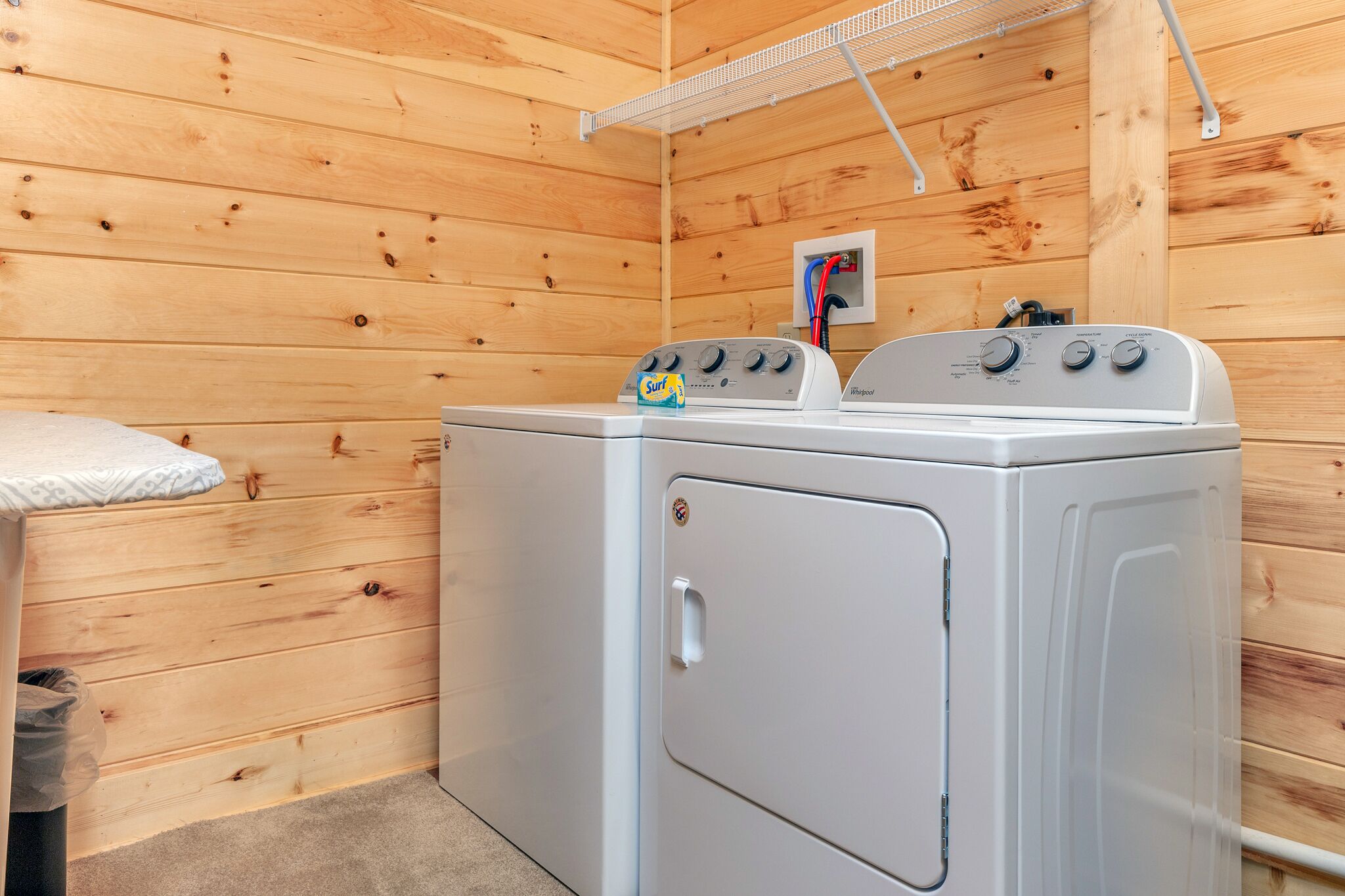 A laundry area with a white washer and dryer, wooden walls, a small shelf with a box, and a power outlet on the wall.