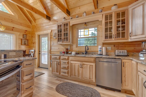 A wooden cabin-style kitchen with granite countertops, wooden cabinets, a dishwasher, and a window with a view of trees.