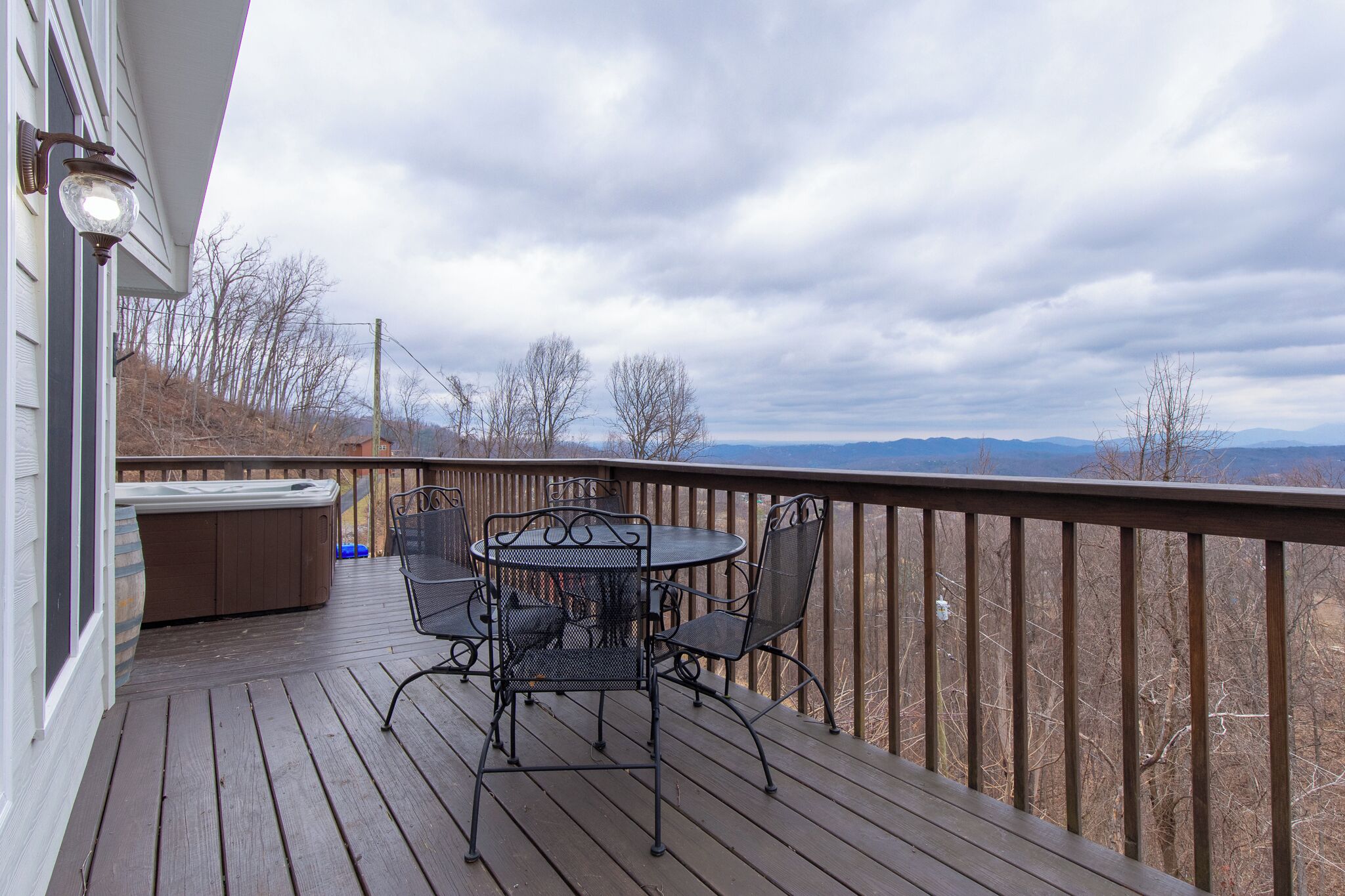 A wooden deck with a hot tub, a table, and chairs, overlooking a mountainous landscape.