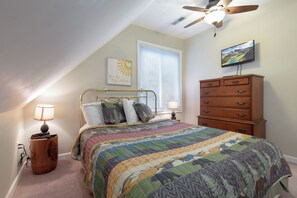Une chambre à coucher comprenant un lit, une commode, un ventilateur de plafond, un téléviseur fixé au mur et une fenêtre avec des stores.