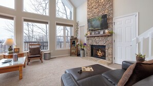 A spacious living room with large windows, a stone fireplace, a flat-screen TV, and a cozy seating area. - Kickback Kabin by Jackson Mountain Rentals (Gatlinburg)
