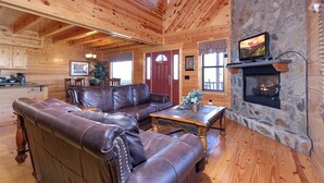 Ein gemütliches Blockhaus-Interieur mit Ledersofa, Kamin aus Stein, Holzwänden und einem darüber montierten Fernseher.