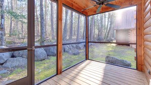 Eine Blockhütte mit großen Fenstern, einem Deckenventilator und Blick auf einen bewaldeten Bereich.