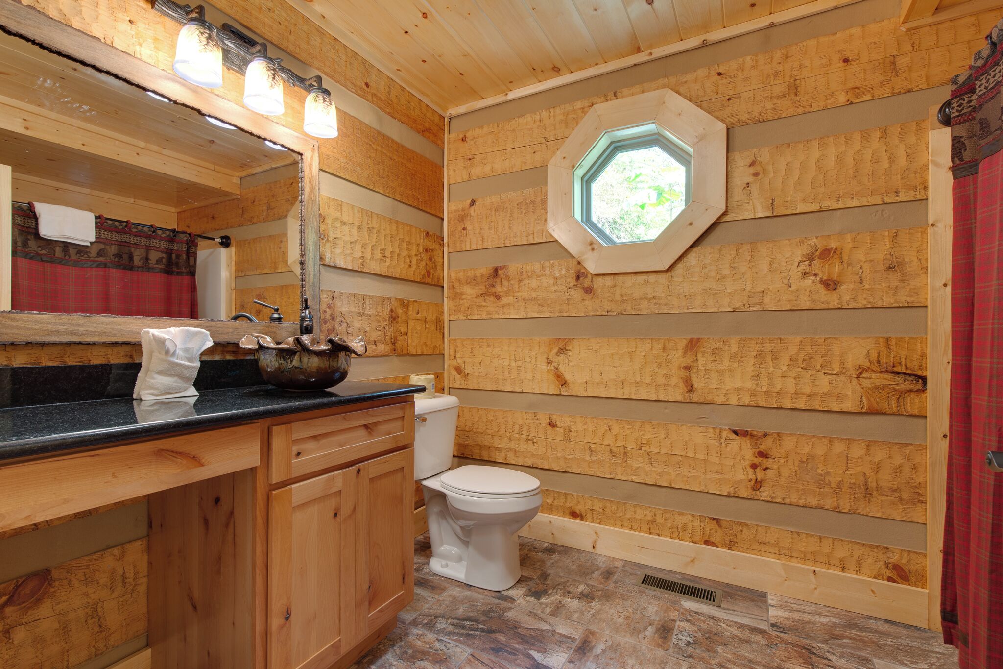Une salle de bain rustique, avec des murs en bois, un plan de toilette noir, des meubles en bois, des toilettes blanches et un puits de lumière.