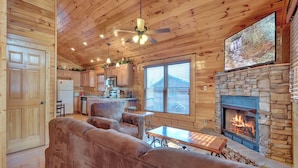 A cozy cabin interior with a stone fireplace, wooden ceiling, and a large TV mounted above it. - Magic Moments II by Jackson Mountain Rentals (Gatlinburg)