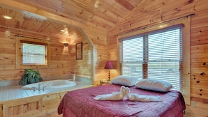 A wooden cabin bedroom with a large bed, a bathtub, and a window with blinds. - Magic Moments II by Jackson Mountain Rentals (Gatlinburg)