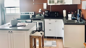 Une cuisine compacte avec des meubles blancs, un plan de travail en bois et un petit coin repas équipé d’un tabouret.