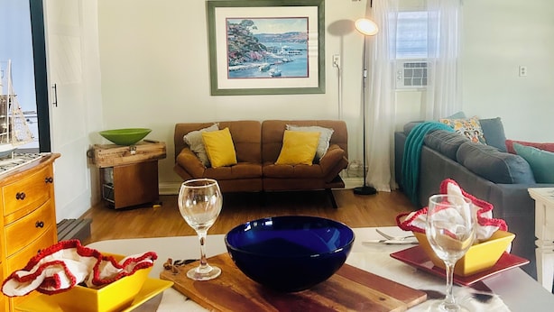Un salon avec un canapé, une table à manger agrémentée d’un saladier bleu et de verres à vin, un tableau encadré au mur et une fenêtre avec des rideaux.