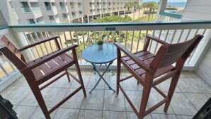 Een balkon met twee houten stoelen en een kleine ronde tafel, met uitzicht op een gebouw met meerdere balkons en een helder zicht op de oceaan.