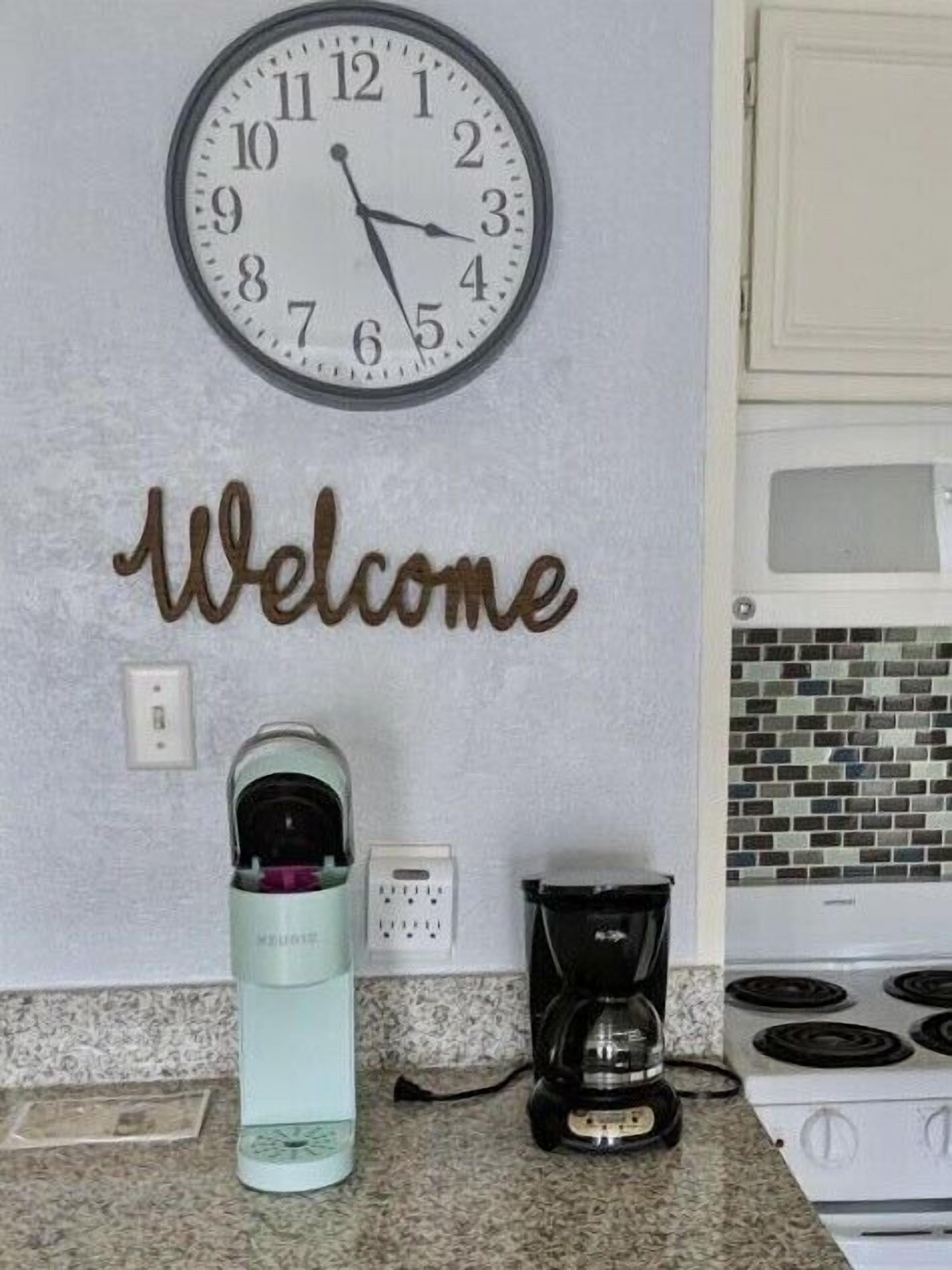 Una cocina con un reloj, un letrero de bienvenida, una tetera, una cafetera y una estufa.