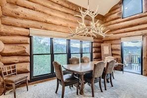 A log cabin interior with a dining table, chairs, a chandelier, and large windows offering a view of the outdoors.