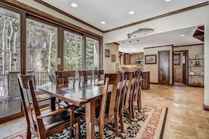 A spacious dining area with a large wooden table and chairs, large windows with a view of trees, and a kitchen in the background. - 🏆 East Vail Home | 4 En-Suite Bedrooms | Heated Garage | Bus to Vail Village (Vail)
