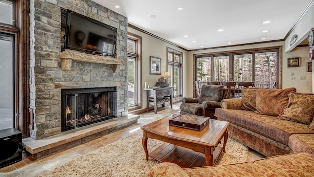 A spacious living room with a stone fireplace, comfortable seating, and large windows offering a view of the outdoors.
