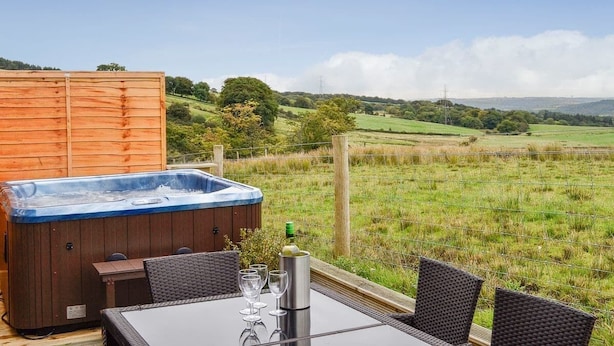 A hot tub with a table and chairs, surrounded by a wooden fence, overlooking a grassy field and hills.