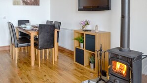 A cozy living room with a wooden dining table, chairs, a TV mounted on the wall, a bookshelf, and a wood-burning stove.