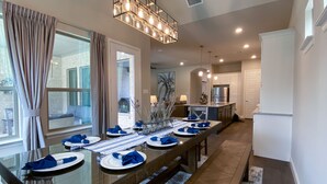 A dining area with a long table set for six, a kitchen in the background, and a view of the outside through large windows. - Entertainment District: Game Room Hot Tub Putting Green 4 Baths Walk to Stadiums (Arlington,)
