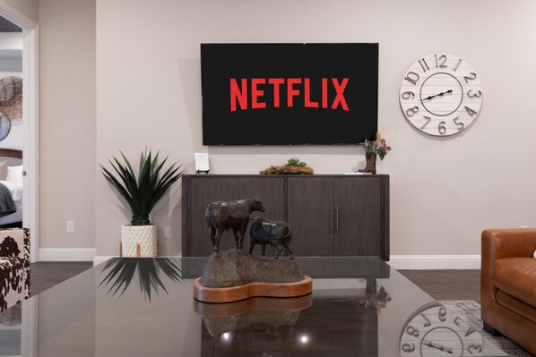 A modern living room with a flat-screen TV displaying "NETFLIX," a decorative clock, a glass coffee table with a cow sculpture, and a brown leather couch.