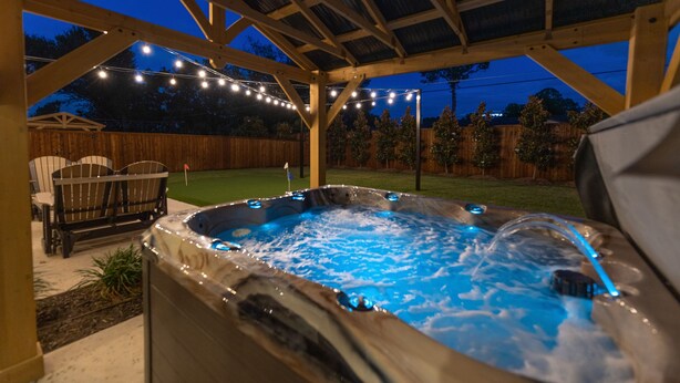 A hot tub under a wooden pergola with string lights, surrounded by a backyard with a lawn and wooden fence.