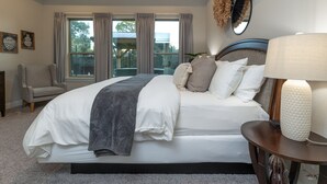 Un dormitorio con una cama grande, una silla, una mesita de noche con lámpara y una ventana con cortinas.