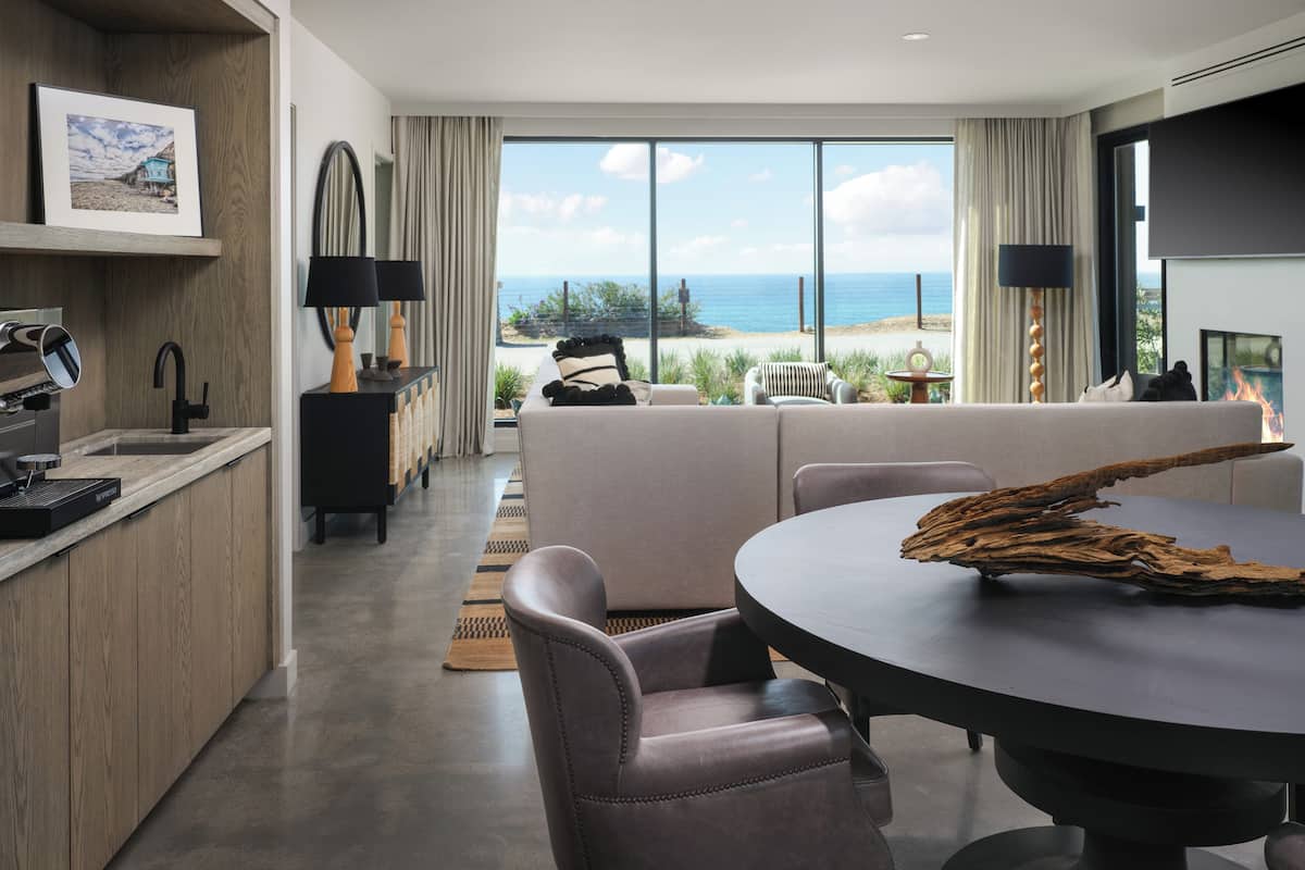 A modern living room with a view of the sea, a dining area, and a kitchen with a coffee machine.